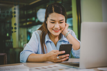 Wall Mural - Business asian woman sitting at her desk with laptop using mobile phone during checking an email or social media on internet.