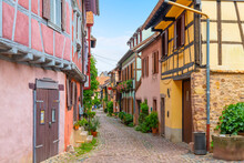 A Narrow Cobblestone Alley Of Colorful Half Timber Buildings In The Medieval Alsatian Village Of Eguisheim, France.