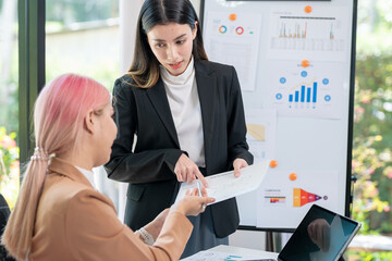 Wall Mural - Two young Asian business women meet to analyze the financial chart at the office to discuss the financial situation at the company. A partner sits at a desk with modern documents and equipment.