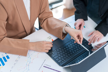 Wall Mural - Two young Asian business women meet to analyze the financial chart at the office to discuss the financial situation at the company. A partner sits at a desk with modern documents and equipment.