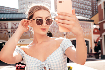 Wall Mural - Young beautiful smiling hipster woman in trendy summer dress clothes. Sexy carefree model posing on the street background at sunset. Positive blond female outdoors. Cheerful and happy. taking selfie