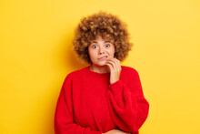 Worried Embarrassed Woman With Oops Expression Looks Scared And Guilty Bites Lips Keeps Fingers On Chin Looks Nervously At Camera Dressed In Red Pullover Isolated Against Yellow Background