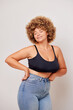 © South House Studio - Sideways shot of self joyed sensual body positive woman with curly hair holds arms on hips feels confident in own body closes eyes and smiles gently wears basic lingerie bra and blue jeans poses over