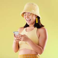 Canvas Print - Phone, laughing and Asian woman typing in studio isolated on a yellow background. Comic emoji, technology and happy female model laugh at funny meme with mobile smartphone for social media or texting