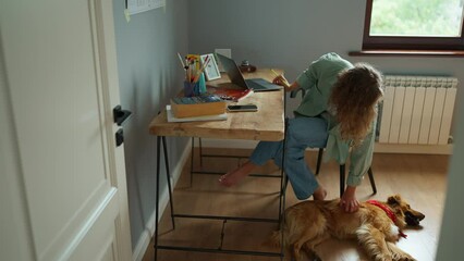 Sticker - Cute curly haired woman working on laptop and stroking her dog lying on the floor at home