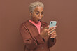 Young shocked beautiful African American woman using mobile phone is dumbfounded by indecent message or photo sent in messenger dressed in casual style posing on isolated brown background.