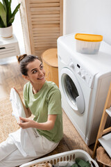 Wall Mural - High angle view of smiling woman looking at box on washing machine near basket with clothes in laundry room.