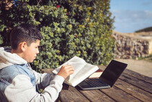 Young Latinstudent Turns The Page Of A Book In Front Of A Laptop, Outdoor
