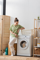 Wall Mural - Young woman looking at laundry in washing machine in laundry room.