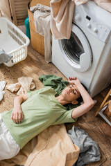 Wall Mural - Top view of tired woman lying around clothes near washing machine in laundry room.