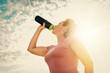 Pretty adult woman in sportswear, drinking from a shaker. Blue sky and clouds in the background. Bottom view. Sunlight. Concept of fitness and sports nutrition
