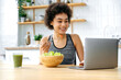 © Kateryna - Positive brazilian or latino curly young woman, sitting at the table in the kitchen, eating a salad, using a laptop, watching a video about healthy eating, recipes. Healthy food, vegetarianism