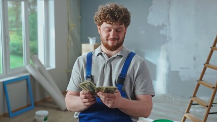 Repairman worker in blue overalls is counting money and smiling. Portrait of a redhead man is posing against backdrop of apartment, ladder, cardboard boxes, window.