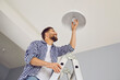 © Studio Romantic - Happy man changing a light bulb at home. High angle shot of a young man in a checkered shirt standing on a step ladder and placing a modern energy-saving lightbulb in a white lamp on the ceiling