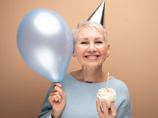 Portrait of her she nice-looking cheery gray-haired lady wearing party hat holding blue balloon and cupcake with candle