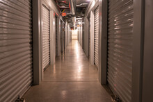 Interior Climate Controlled Rental Storage Units With Exit Signs. Looking Down A Hallway With Locked Metal Roller Doors On Both Sides.