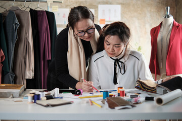 Wall Mural - Asian middle-aged female fashion designer teaches a young teen trainee tailor in studio with colorful thread and sewing fabric for dress design collection ideas, professional boutique small business.