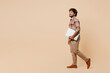 © ViDi Studio - Full body side view young IT Indian man wear brown shirt casual clothes hold closed laptop pc computer walk go isolated on plain pastel light beige background studio portrait People lifestyle concept