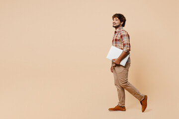 Wall Mural - Full body side view young IT Indian man wear brown shirt casual clothes hold closed laptop pc computer walk go isolated on plain pastel light beige background studio portrait People lifestyle concept