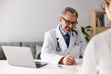 A male doctor sitting with a patient at his modern design office, writing a recipe.