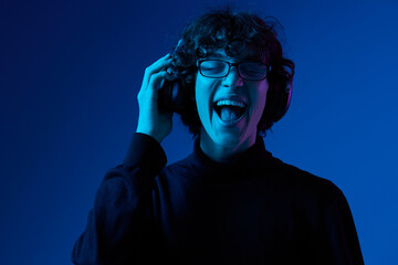Wall Mural - Man teenager wearing headphones listening to music, dancing and singing open mouth smile with glasses, hipster lifestyle, portrait blue background mixed light, fashion style and trends, copying space
