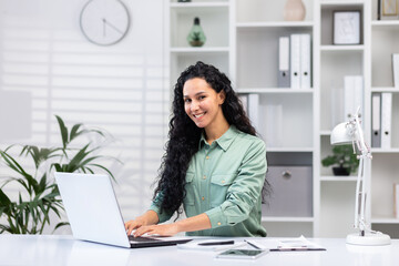 Wall Mural - Young successful hispanic businesswoman working in bright home office indoors sitting at table with documents and reports, woman using laptop at work happy smiling.