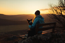 Border Collie Puppy Dog And Young Woman Owner Sitting On A Bench On A Mountain Top At Sunset Seen From Behind