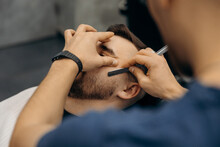 Close Up Photo  Of Trimming A Man's Beard With A Dangerous Razor. A Barber Cuts A Beard In A Barbershop. Beard Trimming In A Beauty Salon For Men