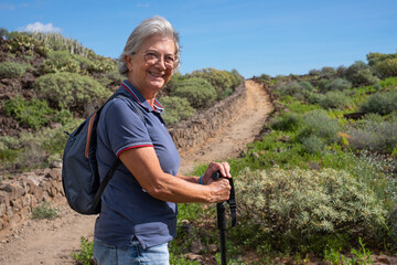 Sticker - Happy senior woman walking in a sunny day on trail outdoors on country excursion. Relaxed caucasian woman enjoying travel or retirement leading healthy lifestyle.