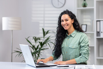 Wall Mural - Portrait of successful beautiful business woman, Hispanic woman in home office smiling and looking at camera, woman at work with laptop typing on keyboard.