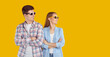 Funny young couple in youth casual clothes and sunglasses looking at copy space orange background. Stylish and cool smiling man and woman with folded hands and smile posing near advertising space.