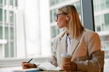 Wall Mural - Busy businesswoman in glasses drinking coffee and making notes while working in cafe