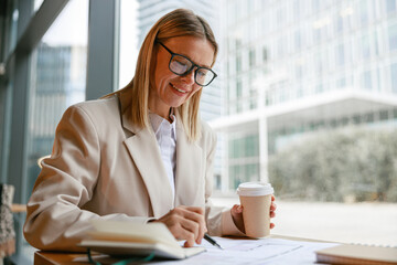 Wall Mural - Smiling businesswoman in glasses drinking coffee and making notes while working in cafe