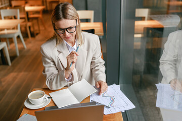 Wall Mural - Focused businesswoman drinking coffee and making notes while working on laptop in cafe
