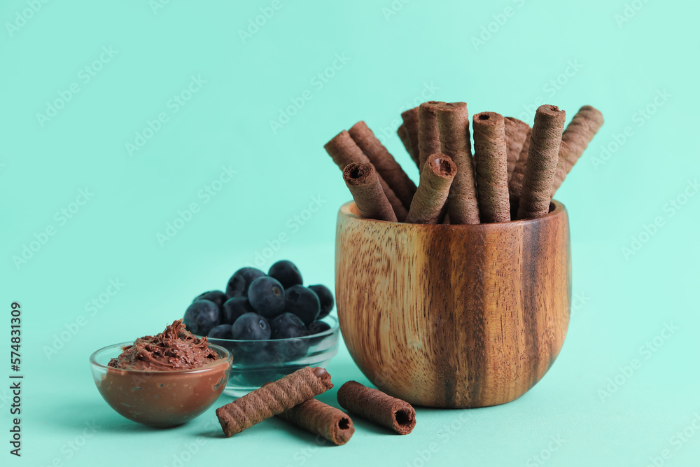 Bowls with delicious chocolate wafer rolls and blueberries on turquoise background