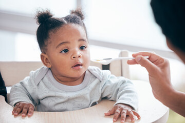 Canvas Print - Food, breakfast and parent feeding a baby in the morning while eating porridge with a mother. Hungry, feed and African child ready to eat a meal, lunch or dinner with a mom in a family home