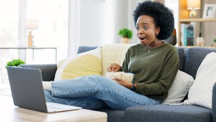 Canvas Print - Happy african american woman streaming online movies on a laptop while snacking on popcorn and relaxing on a sofa at home. Black female enjoying a comedy, eating, laughing and having fun on a weekend