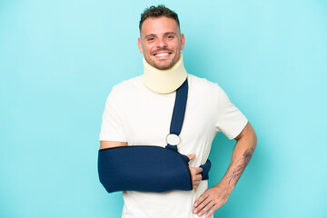 Wall Mural - Young caucasian man wearing a sling  and neck lace isolated on blue background posing with arms at hip and smiling