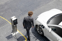 Aerial View Of Progressive Businessman In Black Formal Suit With His Electric Vehicle Recharging Battery At Public Car Park Charging Station As Vehicle Powered By Sustainable Energy Concept.