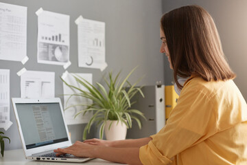 Wall Mural - Side view portrait of woman with brown hair wearing yellow shirt posing in office, sitting at table and typing on keyboard on laptop, doing online project.