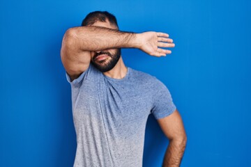 Sticker - Middle east man with beard standing over blue background covering eyes with arm, looking serious and sad. sightless, hiding and rejection concept