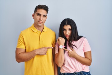 Poster - Young couple standing over isolated background in hurry pointing to watch time, impatience, looking at the camera with relaxed expression