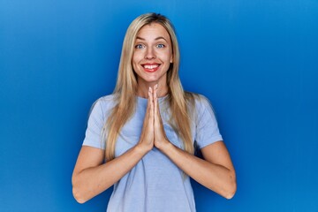 Canvas Print - Beautiful blonde woman wearing casual t shirt over blue background praying with hands together asking for forgiveness smiling confident.