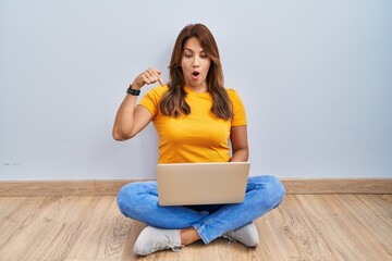 Poster - Hispanic woman using laptop sitting on the floor at home pointing down with fingers showing advertisement, surprised face and open mouth