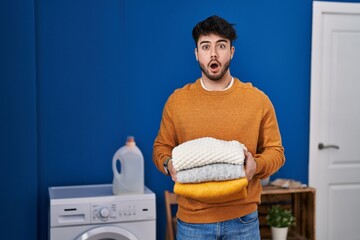 Canvas Print - Hispanic man with beard holding clean folded laundry afraid and shocked with surprise and amazed expression, fear and excited face.