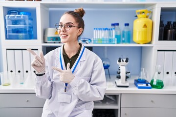 Poster - Young hispanic girl working at scientist laboratory smiling and looking at the camera pointing with two hands and fingers to the side.