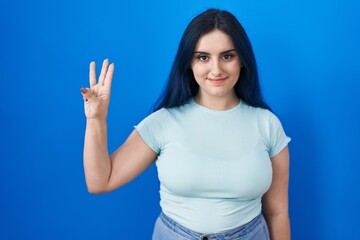 Sticker - Young modern girl with blue hair standing over blue background showing and pointing up with fingers number three while smiling confident and happy.