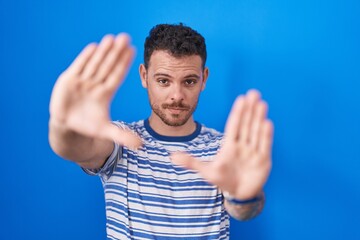 Canvas Print - Young hispanic man standing over blue background doing frame using hands palms and fingers, camera perspective