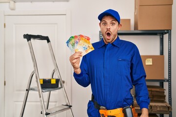 Wall Mural - Young hispanic man working at renovation holding swiss banknotes scared and amazed with open mouth for surprise, disbelief face