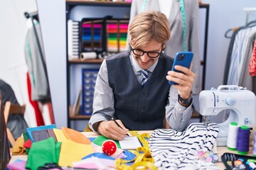 Poster - Young blond man tailor using smartphone writing on notebook at clothing factory